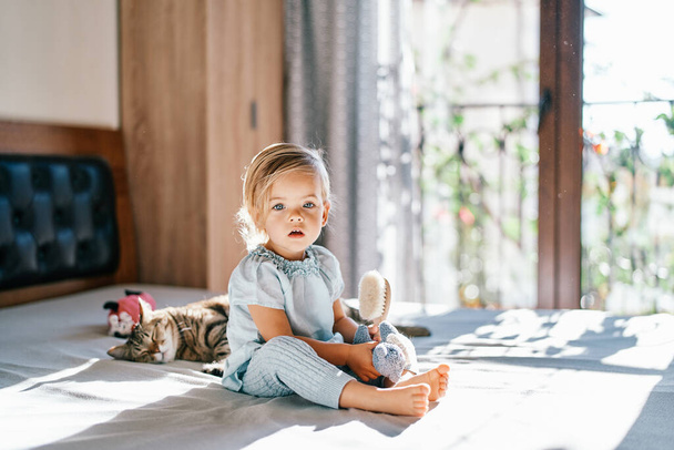 Маленькая девочка с расчёской и игрушкой в руках сидит на кровати рядом со спящей табби-кошкой. Высокое качество фото - Фото, изображение