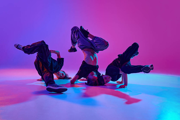 Performance de dança urbana. Desempenho de dança de alta energia por três meninos em luz de néon mista contra fundo de gradiente vibrante. Conceito de esporte e hobby, música, moda e arte, movimento. Anúncio - Foto, Imagem