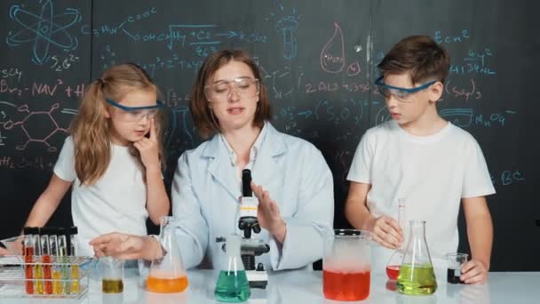 Blanke jongen mengen chemische vloeistof, terwijl de leraar geeft advies. Professionele instructeur dragen lab pak op zoek naar diverse student aan tafel met beker gevuld met gekleurde oplossing. Erutie. - Video