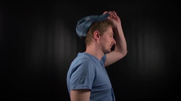 Stylowy młody mężczyzna zakłada czapkę, w niebieskiej koszulce z luźnym zarostem i swobodnym wyglądem, jego przeszywające spojrzenie emanuje ostrą, ale pewną siebie postawą na minimalnym czarnym tle. - Materiał filmowy, wideo