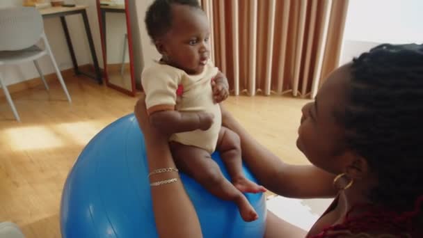 Disparo de mano de madre negra mirando a la hija bebé en la pelota de ejercicio mientras hace ejercicio en casa - Metraje, vídeo