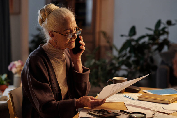 Старша жінка з білим волоссям сидить за столом з фінансовими рахунками і розмовляє з працівником житлового сервісу на мобільному телефоні - Фото, зображення
