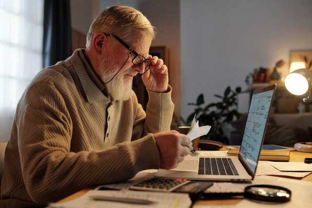 Vue latérale d'un vieil homme sérieux portant des lunettes vérifiant les informations de paiement sur les reçus lorsqu'il est assis devant un ordinateur portable - Photo, image