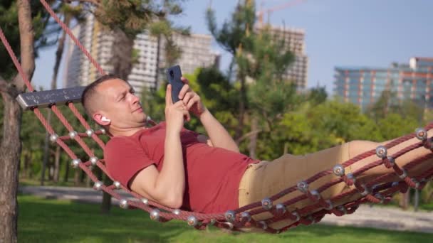 Καυκάσιος νεαρός άνδρας που χρησιμοποιεί smartphone, ακούει μουσική με ακουστικά, σε αιώρα στον κήπο, χαλαρωτικό και καλοκαιρινό concept χρησιμοποιώντας internet ή social media και περιηγείται σε mobile app. - Πλάνα, βίντεο