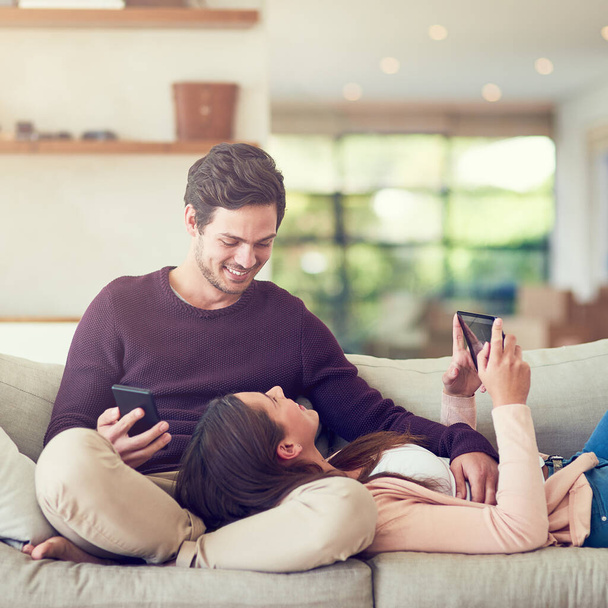 Tablet, telefon komórkowy i para relaksujące się w salonie dla rozrywki razem na kanapie w domu. Happy, miłość i ludzie online networking na telefon i technologii cyfrowej w salonie w mieszkaniu - Zdjęcie, obraz