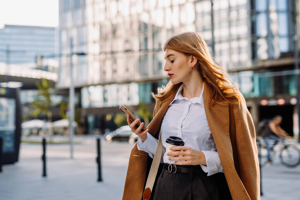 Young positive businesswoman using mobile phone while standing on sunny street near office building, chatting online with friends during break at work - Photo, Image
