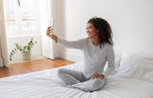 ヒスパニック系の女性がベッドの上に座り,自撮りをするためにスマートフォンを腕の長さで握った. 彼女はスクリーンに焦点を当て,ベッドに快適に座っている間完璧なショットを確保します. - 写真・画像