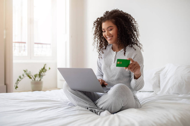 Mulher hispânica está sentada em uma cama, segurando um cartão de crédito e um laptop em suas mãos. Ela parece estar envolvida em compras online ou transações financeiras.. - Foto, Imagem