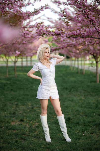 Una hermosa rubia con un vestido blanco se encuentra en un parque sobre el fondo de los árboles florecientes. Foto de alta calidad - Foto, imagen