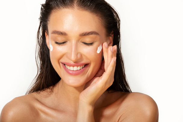 Concepto de cuidado de la piel. Mujer europea feliz aplicando crema hidratante cosmética en su cara y sonriendo con los ojos cerrados sobre fondo blanco - Foto, Imagen