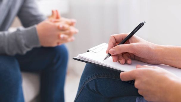 Ένα κοντινό πλάνο που δείχνει ένα θεραπευτή χέρια κρατώντας ένα στυλό και γράφοντας σε ένα σημειωματάριο, με τους ασθενείς χέρια στο παρασκήνιο gesturing μέση της συζήτησης, περικοπεί - Φωτογραφία, εικόνα