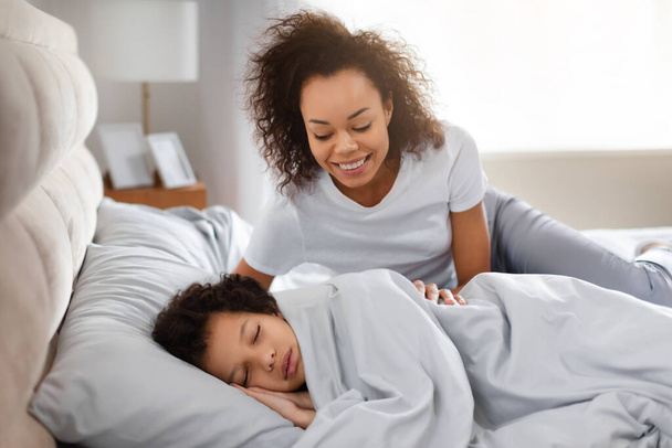 Africká americká matka se něžně usmívá na své malé dítě, které ještě spí, zabalené v útulném ložním prádle. Měkké ranní světlo naplňuje místnost teplem a naznačuje začátek nového dne.. - Fotografie, Obrázek