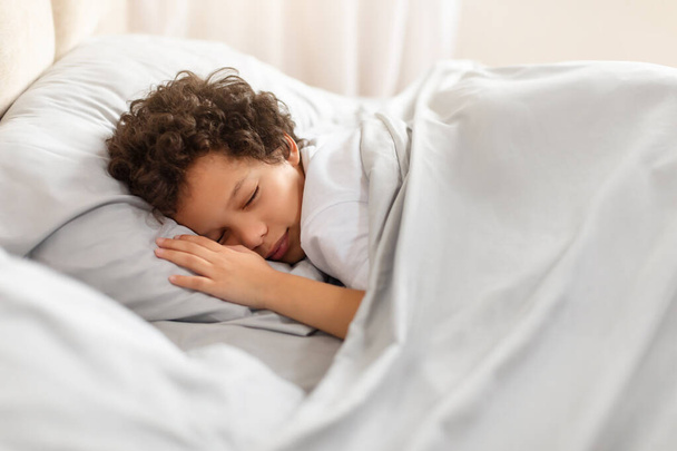 Um jovem afro-americano está dormindo em paz em uma cama com lençóis brancos crocantes cobrindo-o. Sua cabeça repousa sobre um travesseiro fofo enquanto ele encontra descanso e conforto em seu entorno. - Foto, Imagem
