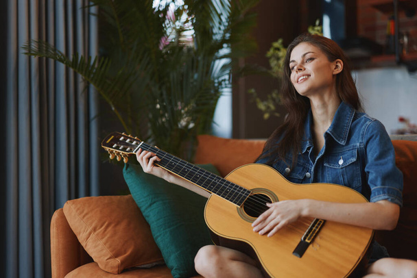 自宅のインテリアでリビングルームのソファーでアコースティックギターを演奏する若い女性 - 写真・画像