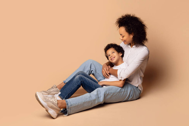 A jovem mãe afro-americana senta-se no chão, embalando seu filho em seus braços com um abraço caloroso. O fundo é um bege liso, macio - Foto, Imagem