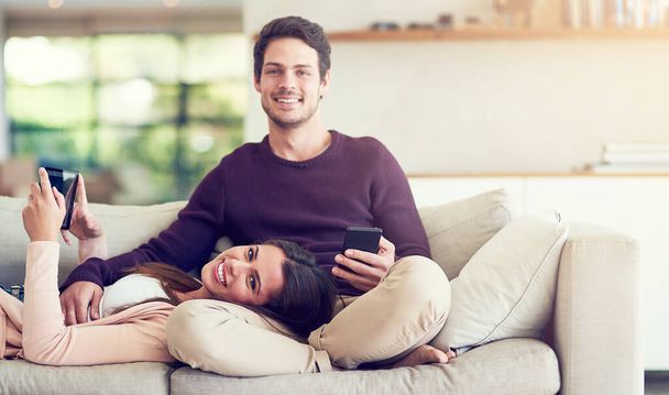 Couple, portrait and relax on sofa with phone, tablet and connectivity for app in home living room. Man, woman and technology with bonding, care and morning on vacation with smile on couch in house. - Photo, Image