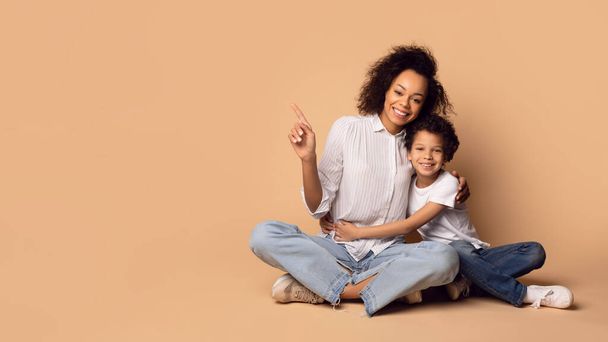 Μια χαρούμενη Αφρο-Αμερικανίδα μητέρα και ο νεαρός γιος της κάθονται στο πάτωμα με τις πλάτες τους σε ένα απαλό μπεζ φόντο, δείχνοντας και κοιτάζοντας το χώρο των αντιγράφων. - Φωτογραφία, εικόνα