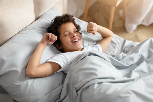 Афроамериканський хлопчик зручно лежить у ліжку, з м'якою посмішкою на обличчі. Він виглядає задоволеним і розслабленим, насолоджуючись миттю спокою перед сном, розтягуючись - Фото, зображення