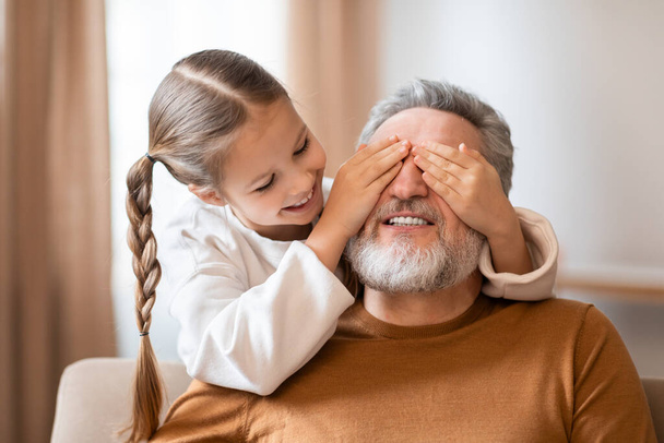 Молодая девушка с хвостиком закрывает дедушкины глаза сзади, когда они сидят вместе, создавая момент игривой связи. Дедушка с закрытыми глазами смеется. - Фото, изображение
