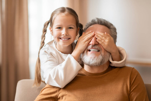 勇敢な髪を持つ若い女の子が後ろから祖父の目を覆い,暖かく照らされた部屋で笑いを分かち合い,遊び心のあるゲームを示唆しています - 写真・画像