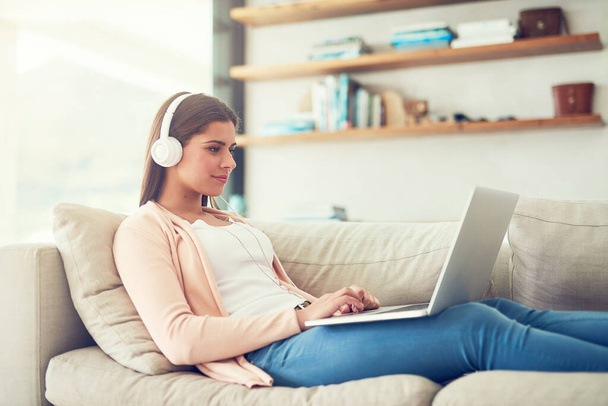Woman, laptop and headphones with video on sofa in home for audio streaming, subscription service and social media. Person, music and watching series with movie entertainment, radio playlist or sound. - Photo, Image