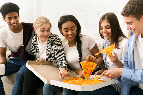 Группа друзей, сидящих на диване и наслаждающихся кусочками пиццы. Разнообразная группа вступает в непринужденную беседу, наслаждаясь обедом вместе. - Фото, изображение