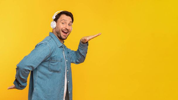 Um homem sorrindo usando fones de ouvido e uma jaqueta de ganga está contra um cenário amarelo vibrante, ouvindo música e dança, espaço de cópia - Foto, Imagem