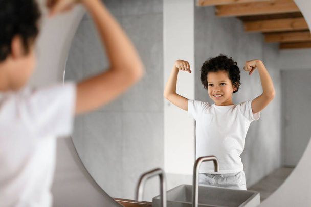 Criança afro-americana está de pé na frente de um espelho do banheiro, orgulhosamente flexionando seus músculos e exibindo um sorriso alegre. O banheiro tem um design moderno com vigas de madeira visíveis - Foto, Imagem