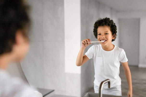 O jovem afro-americano está de pé em frente a um espelho, lavando diligentemente os dentes. Ele está focado em seu reflexo, segurando uma escova de dentes e pasta de dentes - Foto, Imagem