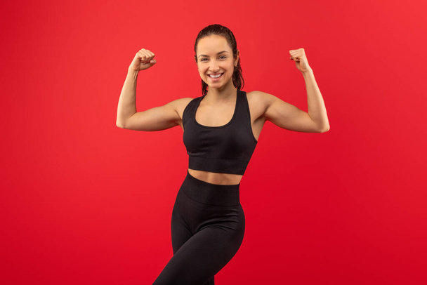 Eine junge Frau steht selbstbewusst mit gebeugten Armen da, um ihre Muskeln zur Schau zu stellen, trägt einen schwarzen Sport-BH und Leggings, alles vor einem leuchtend roten Hintergrund. - Foto, Bild