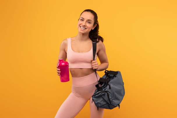 Egy vidám fiatal nő rózsaszín sportmelltartóban és lábszárvédőben magabiztosan áll egy élénk sárga háttér előtt, egyik kezében egy vizes palackkal, a vállán pedig egy sporttáskával. - Fotó, kép