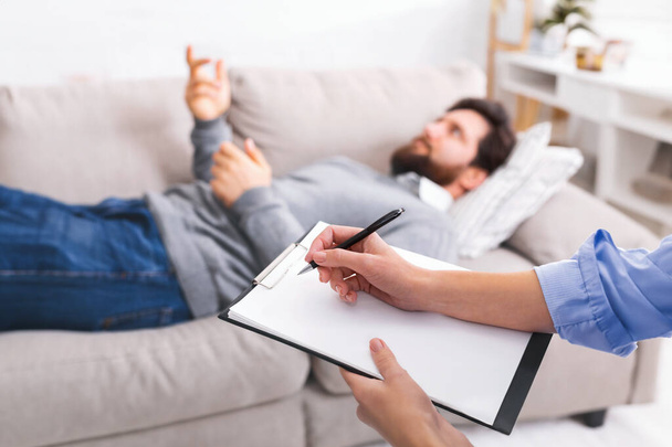 A man is lying on a couch, gesturing with his hand as he speaks, while engaged in a counseling session. A therapist, partially visible in the foreground, is attentively taking notes on a clipboard. - Photo, Image