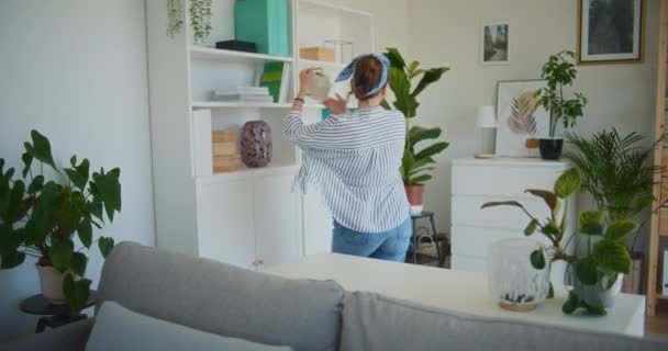 Eine Frau hört Musik, tanzt und gießt Blumen in ihrem Wohnzimmer und genießt einen lebhaften Moment - Filmmaterial, Video