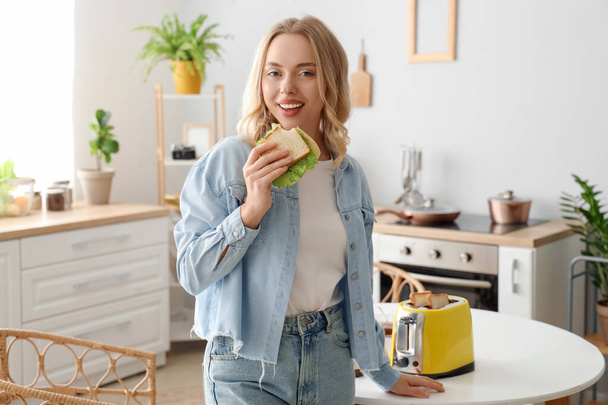 Young beautiful woman eating tasty sandwich in kitchen - Photo, Image