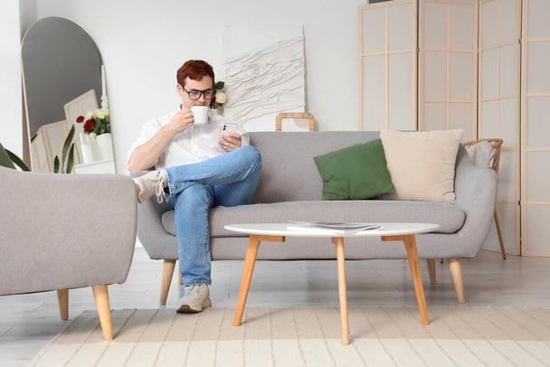 Νεαρός άνδρας με γυαλιά ηλίου με φλιτζάνι τσάι και κινητό τηλέφωνο κάθεται σε γκρι καναπέ στο σπίτι - Φωτογραφία, εικόνα