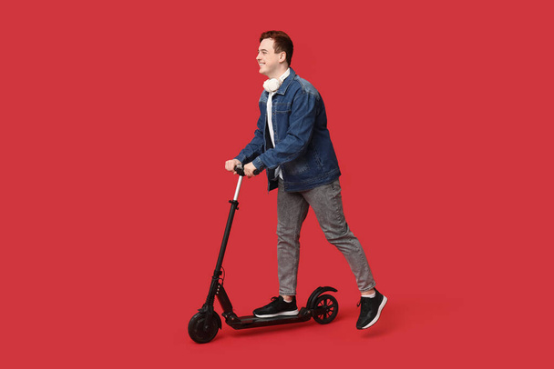 Joven montando moderno scooter de patada eléctrica con auriculares sobre fondo rojo - Foto, Imagen