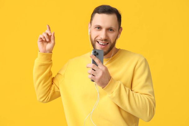 Hombre joven con auriculares modernos con teléfono móvil apuntando a algo en el fondo amarillo - Foto, Imagen