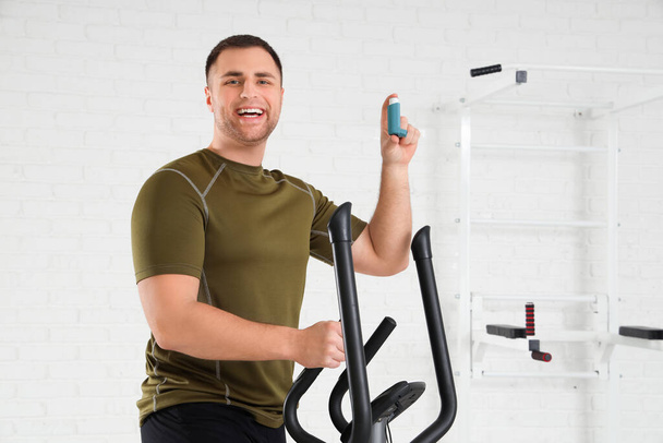 Sportlicher junger Mann mit Inhalator auf Heimtrainer im Fitnessstudio - Foto, Bild