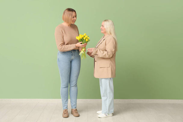Взрослая женщина приветствует свою мать букетом тюльпанов у зеленой стены. Международный женский день - Фото, изображение