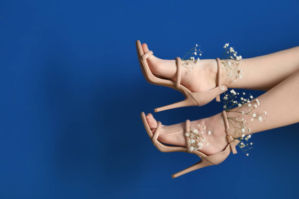 Ноги красивої молодої жінки в бежевих щиколотках взуття з білими квітами гіпсофіли на синьому фоні - Фото, зображення