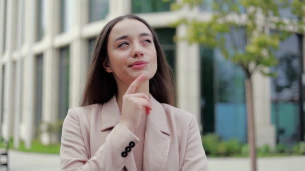 Pensive woman in stylish jacket touching chin with index finger while pursing lips and looking aside. Playful designer checking exterior of modern buildings and creating new renovating ideas in mind. - Footage, Video