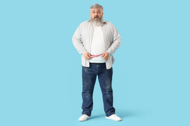 Overgewicht volwassen geschokte man met meetlint op blauwe achtergrond. Gewichtsverlies concept - Foto, afbeelding