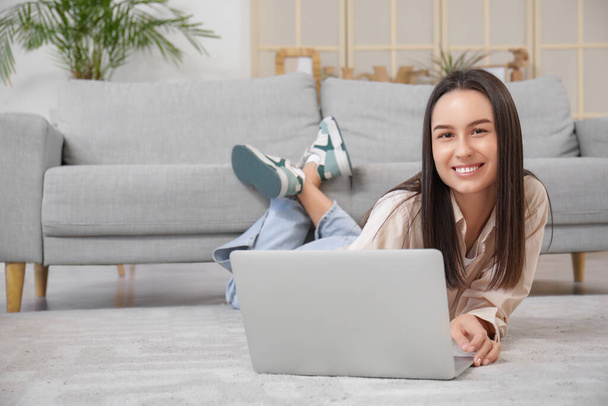 Красива молода жінка з ноутбуком лежить на підлозі біля сірого дивана у вітальні - Фото, зображення