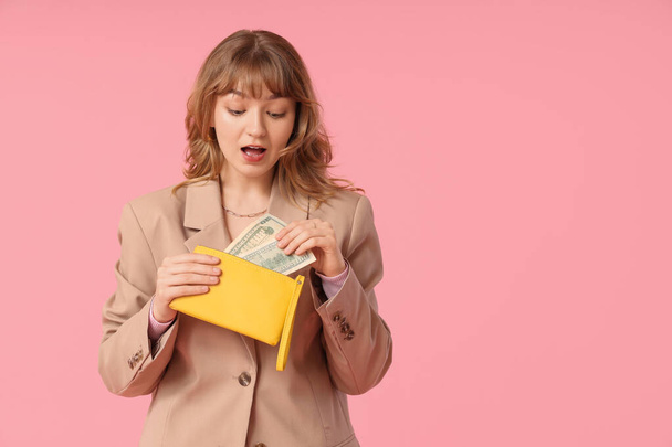Έκπληκτη νεαρή γυναίκα κρατώντας κίτρινο πορτοφόλι και τα χρήματα σε ροζ φόντο - Φωτογραφία, εικόνα