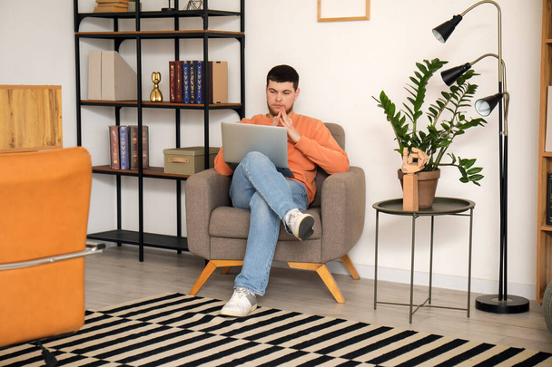 Молодой бородатый мужчина использует ноутбук в кресле в офисе - Фото, изображение