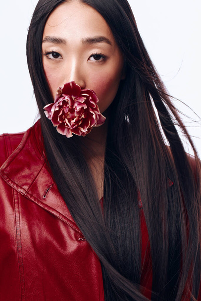 Ελκυστική νεαρή γυναίκα με μακριά μαύρα μαλλιά και κόκκινο δερμάτινο μπουφάν, κρατώντας ένα λουλούδι στο στόμα - Φωτογραφία, εικόνα