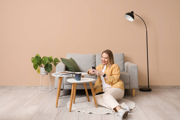 Mujer joven usando el teléfono móvil y sentada en el suelo en la sala de estar beige - Foto, imagen