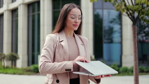 Portrait d'une femme joyeuse fermant ordinateur portable personnel tout en posant sur fond de bâtiment contemporain. Positif jeune femme caucasienne portant des lunettes regardant la caméra avec un sourire agréable. - Séquence, vidéo