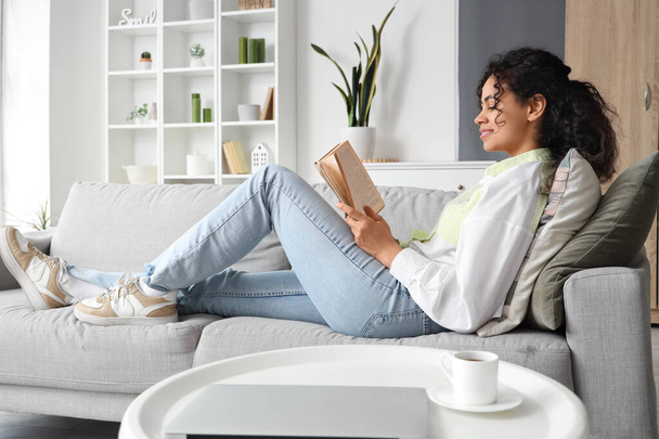 Fiatal afro-amerikai nő olvassa könyvet kanapén otthon - Fotó, kép
