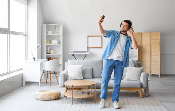 Молодой бородатый мужчина в наушниках с мобильным телефоном слушает музыку дома - Фото, изображение